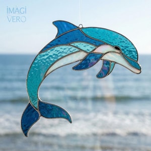 以下が含まれることがあります： 青と白の色合いのステンドグラスのイルカ。左上に「IMAGI VERO」の文字があります。イルカはぼやけた海の背景の前に吊り下げられています。ガラスは質感のある外観をしています。