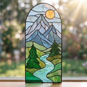 May include: An arched stained glass panel featuring a landscape scene with a yellow sun, blue and white mountains, green pine trees, and a winding light blue river. The glass pieces are joined by dark metal lines, creating a detailed nature motif.