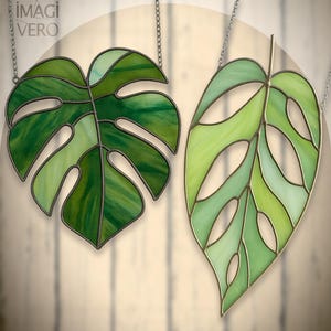 May include: Two stained glass leaf pendants, one a split-leaf philodendron and the other a stylized leaf, hang from silver chains. The leaves feature shades of green and are outlined in a dark metal. The brand name "IMAGI VERO" is in the upper left corner.