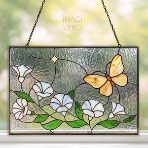 Puede incluir: Panel rectangular de vidrieras con una mariposa y flores blancas con hojas verdes. El panel está enmarcado en metal marrón y cuelga de una cadena. El texto "iMAGI VERO" es visible.