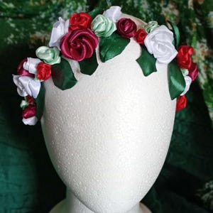 May include: A floral crown featuring red, white, and green satin ribbon roses and leaves. The crown is displayed on a white mannequin head. The background is a dark green fabric.