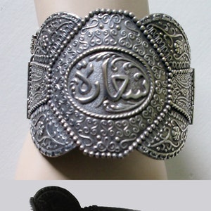 May include: A silver bracelet with an ornate design and a hinged clasp. The bracelet features a large oval centrepiece with an inscription in Arabic script.