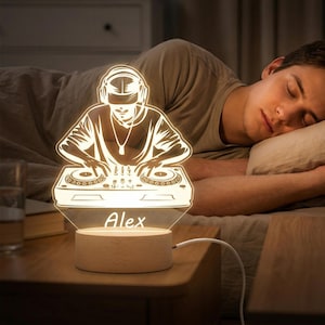 May include: A glowing LED night light featuring a DJ design with the name "Alex" etched on the base. The light is set on a wooden base and plugged into a wall outlet. The image shows a person sleeping in a bed next to the lamp.