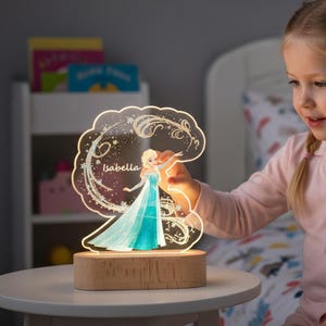 May include: A personalized night light featuring a princess in a blue dress, with the name "Isabella" on the front. The light is set on a wooden base and emits a warm glow. The design includes decorative swirls and stars.