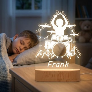 Op de afbeelding: Gepersonaliseerde nachtlamp met een drummer silhouet, drumstel en de naam "Frank" in het wit. De lamp heeft een houten voet en is aangesloten. De achtergrond toont een slapend kind in een slaapkamer.