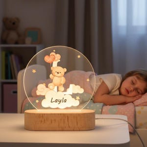 May include: A personalized night light featuring a teddy bear holding heart-shaped balloons, clouds, and stars. The name "Layla" is displayed on the clouds. The light sits on a wooden base and is plugged in.