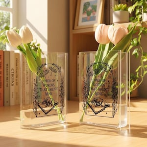 Pode incluir: Dois vasos retangulares transparentes com o texto "LOVER OF WORLDS THAT AREN'T MINE" e uma ilustração de livro. Cada vaso contém água e algumas tulipas. Os vasos estão sobre uma superfície de madeira, com livros e plantas ao fundo.