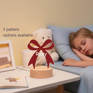 Op de afbeelding: Een rood strikvormig nachtlampje met de witte naam "Riley" op een houten voet. De tekst "3 patroonopties beschikbaar" is zichtbaar. Op de nachtkastje liggen een teddybeer en een slapend kind.