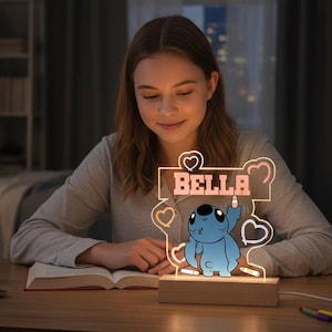 May include: A personalised night light with a blue cartoon character and the name "BELLA" in pink. The design is on clear acrylic with heart shapes, set on a wooden base. The light is on a wooden table.