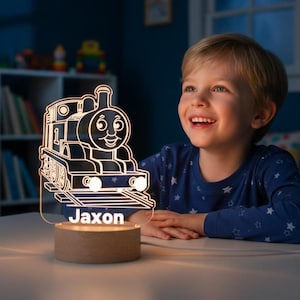 Könnte beinhalten: Ein nachtleuchtendes Nachtlicht in Form einer Lokomotive mit dem Namen "Jaxon" auf der Vorderseite. Das Licht besteht aus klarem Acryl und steht auf einem Holzsockel. Das Design der Lokomotive zeigt ein lächelndes Gesicht und wird von einem weichen, warmen Licht beleuchtet.