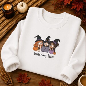 May include: A white crewneck sweatshirt featuring a graphic of three cartoon characters wearing black witch hats and looking at a laptop. The text "Witching Hour" is printed below the illustration, which includes orange and purple pumpkins.