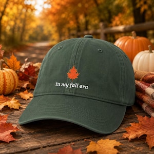 May include: A dark green baseball cap featuring an embroidered orange maple leaf centred above the white text "in my fall era". The cap has a curved brim, a structured crown with eyelets, and a button at the top, showcasing a classic design.