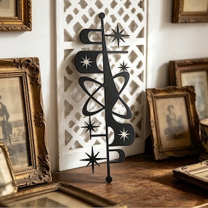 May include: A black metal wall art piece with a mid-century modern design. The art features starburst and atomic shapes, with a central vertical element and various curved and circular forms. The piece is displayed against a white background and surrounded by framed pictures.