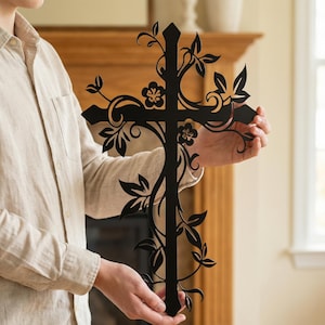 May include: A black metal cross with a floral vine design. The cross features cut-out leaves and flowers, creating a decorative and elegant appearance. The cross is held in front of a neutral background.