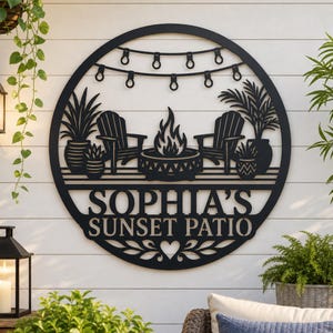 May include: A circular black metal wall sign featuring a cutout design of two chairs, a fire pit, potted plants, and string lights. The text "SOPHIA'S SUNSET PATIO" is centered in the lower half, accented by a heart and leaf motifs at the bottom.