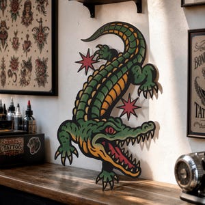May include: A wall-mounted cutout of a green and yellow alligator with a red eye and open mouth showing sharp teeth. The alligator is depicted in a traditional tattoo art style with bold black outlines and two red eight-pointed star accents.