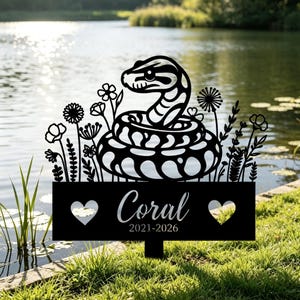 May include: Black metal garden stake featuring a stylized snake design with floral accents. The stake includes the name "Coral" and the years "2021-2026" with heart-shaped cutouts. The stake is placed in a grassy area near a body of water.