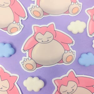 May include: Pink and beige Snorlax stickers with a smiling expression, surrounded by white, blue, and pink cloud-shaped embellishments on a purple background. The stickers are in a cartoon style.