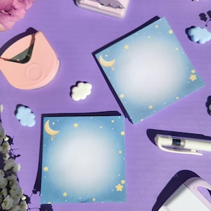 Pode incluir: Uma composição de artigos de papelaria sobre uma superfície roxa. Dois blocos de notas quadrados com fundo gradiente azul, ilustrações de lua e estrelas e um centro branco estão presentes. Outros itens incluem um arredondador de cantos rosa, um furador rosa e enfeites em forma de nuvem.