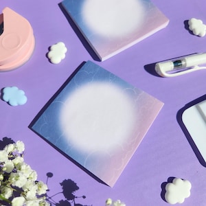 May include: Two square sticky note pads with a blue and pink gradient design and a white centre. The pads are surrounded by white cloud-shaped decorations and a pen on a purple surface. A sprig of white flowers is in the corner.