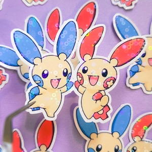 May include: A collection of die-cut stickers featuring a cartoon creature with a light yellow body, large ears colored in red and blue, and a happy expression. The stickers have a holographic finish with a circular pattern that reflects light.