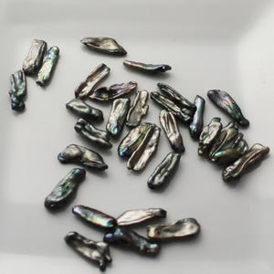 May include: A collection of iridescent black and green freshwater pearls, each with a unique shape and size. The pearls are arranged on a white surface.
