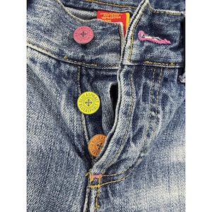 Puede incluir: Vaqueros de mezclilla azul con cierre de tres botones distintos en rosa, amarillo y naranja, cada uno grabado con el texto Blue Asphalt. Los vaqueros presentan un lavado desgastado, costuras doradas y una etiqueta interior roja con texto.
