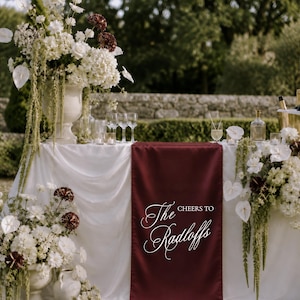 Op de afbeelding: Een wit gedrapeerde tafel met een diepbordeauxrode tafelloper met de witte tekst "CHEERS TO The Radloffs". De tafel is versierd met grote bloemstukken van witte en donkerrode bloemen, helder glaswerk en flessen.