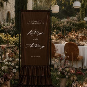 May include: A coffee brown silk satin wedding welcome sign featuring the text "WELCOME TO THE WEDDING OF Kathryn AND Anthony 10.10.2026" in white lettering. The rectangular fabric banner is mounted on a black frame and has tiered ruffles.