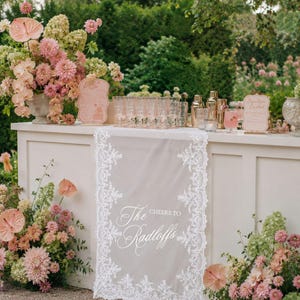 Könnte beinhalten: Ein weißer Tischläufer mit aufwendigen Spitzenrändern und dem mittig aufgedruckten weißen Text "Cheers to The Radloffs". Der Läufer liegt auf einer weißen Bartheke, die mit Blumenarrangements und verschiedenen Gläsern dekoriert ist.