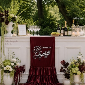 May include: A burgundy silk satin bar runner featuring a tiered ruffled bottom and white script text that reads Cheers to The Radloffs. The runner is draped over a white bar counter decorated with floral arrangements, wine bottles, and glassware.