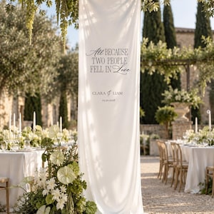 Peut inclure: Une longue bannière en tissu blanc vertical avec le texte imprimé en noir : All BECAUSE TWO PEOPLE FELL IN Love, CLARA & LIAM, 05.20.2028. La bannière est suspendue à une structure et repose sur le sol, entourée de compositions florales vertes.
