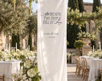 May include: A tall, vertical white fabric banner featuring black printed text that reads: All BECAUSE TWO PEOPLE FELL IN Love, CLARA & LIAM, 05.20.2028. The banner hangs from a structure and pools slightly at the base, surrounded by green floral arrangements.