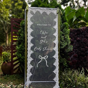 May include: A rectangular white metal frame holding a sheer white fabric panel with intricate lace borders. The fabric features the white printed text WELCOME TO Chloe and Steh's Baby Shower and a white line drawing of a bow tied with ribbon.