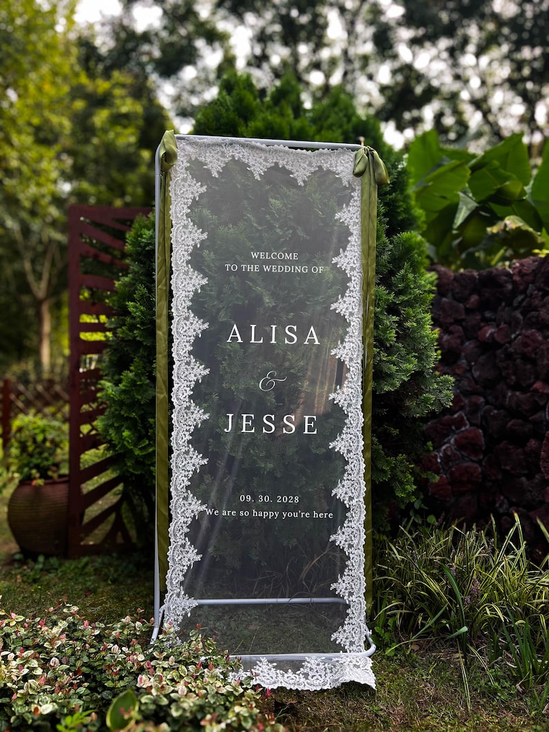 May include: A rectangular clear acrylic wedding sign held in a white metal frame, draped with white lace trim along the vertical edges and olive green ribbons at the top. The text reads: WELCOME TO THE WEDDING OF ALISA & JESSE 09. 30. 2028 We are so happy you're here.