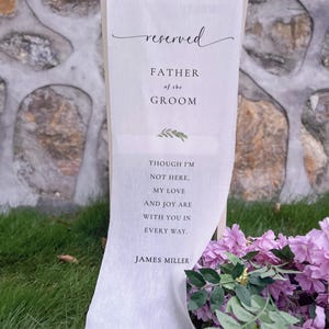 Könnte beinhalten: Ein weißes Stoffschild, das über einem Stuhl drapiert ist, mit dem Wort "reserved" in eleganter Schrift. Das Schild lautet "FATHER of the GROOM" und enthält eine herzliche Botschaft und den Namen "JAMES MILLER". Violette Blumen sind im Vordergrund.