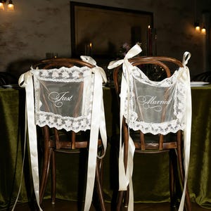 May include: Two wooden chairs decorated for a wedding. Each chair has a white lace cover with the words "Just" and "Married" embroidered on them. Long, ivory ribbons are tied to the chairs.