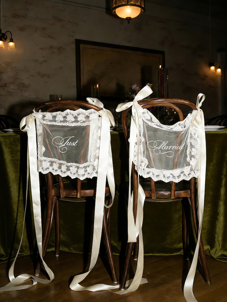 May include: Two wooden chairs decorated for a wedding. Each chair has a white lace cover with the words "Just" and "Married" embroidered on them. Long, ivory ribbons are tied to the chairs.