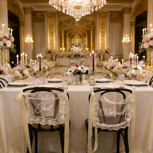 Op de afbeelding: Elegante bruiloftsreceptie setting met een lange tafel bedekt met een wit tafelkleed. Twee stoelen met transparante ruggen met de tekst "Just" en "Married" staan op de voorgrond. De kamer is versierd met bloemen, kaarsen en een grote kroonluchter.