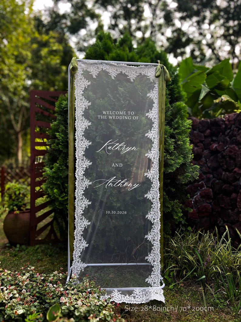 May include: A rectangular wedding welcome sign featuring a clear acrylic panel framed by white lace trim and olive green ribbons. The text reads: WELCOME TO THE WEDDING OF Kathryn AND Anthony 10.10.2026. The sign measures 70 by 200 cm.