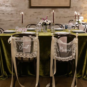 May include: Wedding reception table setting with a green velvet tablecloth, candles, and floral arrangements. Two chairs are decorated with lace and ribbons, with the words "Just" and "Married" displayed.