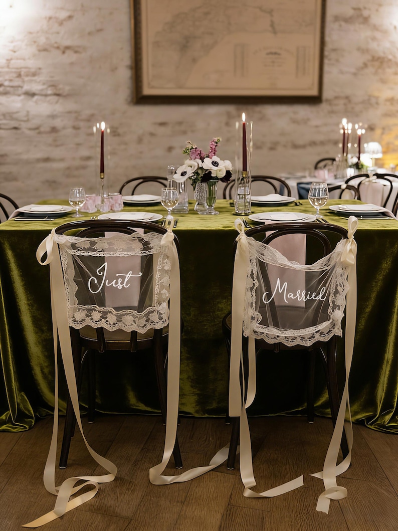 May include: Wedding reception table setting with a green velvet tablecloth, candles, and floral arrangements. Two chairs are decorated with lace and ribbons, with the words "Just" and "Married" displayed.