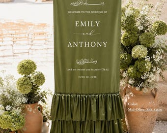 May include: A tall, rectangular mid-olive silk satin wedding welcome sign featuring white text that reads: Welcome to the wedding of Emily and Anthony, and we created you in pairs [78:8], June 10, 2028. The base has three tiers of ruffles.