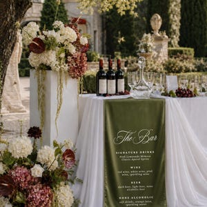 Può includere: Un tavolo bianco coperto da una tovaglia bianca e un runner verde oliva con il testo The Bar, Signature Drinks, Wine, Beer e None Alcoholic. Tre bottiglie di vino, un decanter in vetro e dei bicchieri sono disposti sul tavolo.