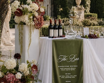 Peut inclure: Une table de bar blanche recouverte d'une nappe blanche et d'un chemin de table en velours vert olive portant le texte The Bar, Signature Drinks, Wine, Beer et None Alcoholic. Trois bouteilles de vin, une carafe et des verres sont posés.