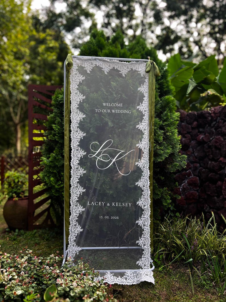 May include: A rectangular wedding welcome sign featuring a sheer white fabric panel with intricate white lace trim along the vertical edges. The sign displays the text "WELCOME TO OUR WEDDING", the monogram "LK", "LACEY & KELSEY", and "15. 05. 2026".