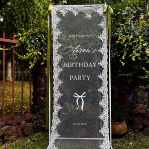 May include: A white framed sign with a sheer, lace-edged panel. The sign reads "Welcome to Aaron's Birthday Party" with a bow graphic and the date 28.09.2026. Green ribbons adorn the top.