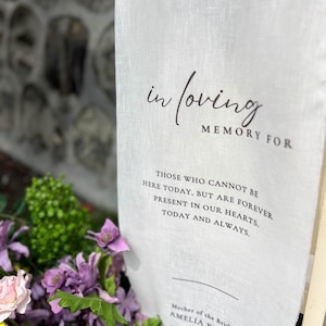 Peut inclure: Un panneau en lin blanc avec les mots "in loving memory for" et un message touchant. Le panneau est entouré de fleurs violettes et roses, avec un feuillage vert. Le texte comprend des noms et des titres.