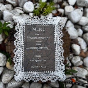 Op de afbeelding: Een rechthoekige menukaart met een witte kanten rand. Het menu bevat het woord "MENU" bovenaan, gevolgd door de gangen voorgerecht, hoofdgerecht en dessert. Het menu bevat de namen van de gerechten en de namen "OLIVIA + ALEX" onderaan.