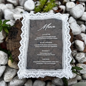 Può includere: Un menu trasparente con bordo in pizzo bianco, che mostra un menu per un pasto. Il menu comprende antipasti, piatti principali, dessert e bevande. La carta è posta su una superficie di legno.